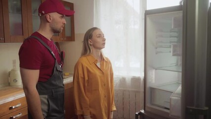 Homeowner explains to a repairman the malfunction of her refrigerator, gesturing and pointing at the appliance while he listens attentively in the kitchen
