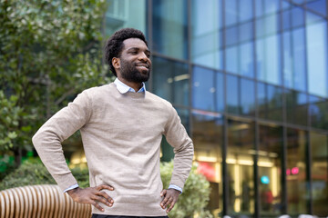 A smiling man with hands on hips poses confidently outside a modern glass building. The soft sunlight enhances the happy expression on his face.