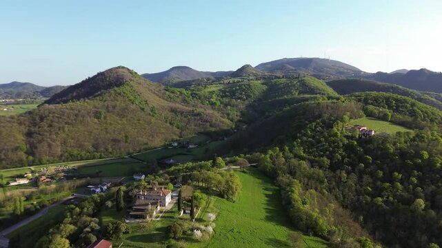 Panning aerial view of an old house in a green landscape with Monte Venda mountain in Colli Euganei hills with setting sun casting warm light on the hillside near rows of grapevines
