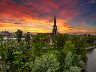Fototapeta premium Aerial view of Frankfurt during sunset, featuring a historic church with a tall spire, lush green trees, a river, and modern city buildings in the background.