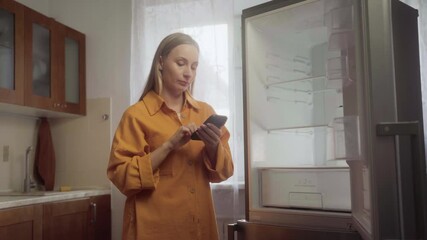 Frustrated female homeowner struggling with malfunctioning refrigerator, using smartphone to contact repair service while experiencing kitchen appliance breakdown