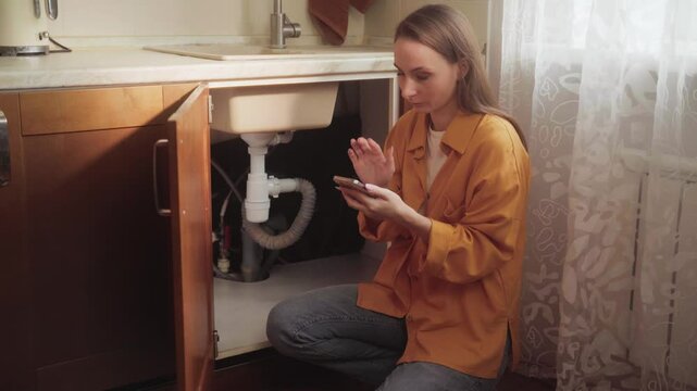 Female homeowner browsing smartphone app for selecting professional plumber, resolving kitchen sink drain clogging issue through convenient online service technology
