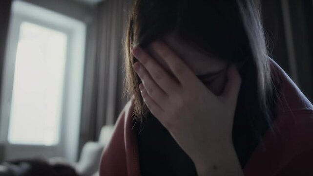 Tilting closeup of teenage schoolgirl sitting at home, suffering doing cyberbullying after reading messages on smartphone, covering face and crying, unable to concentrate on homework