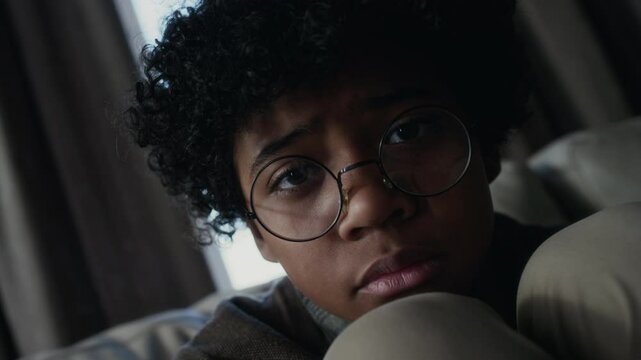 Close-up Dutch angle portrait of face of geeky young African American boy sitting hunched up on couch, looking at camera with sad tearful eyes, worried about online bullying and loneliness