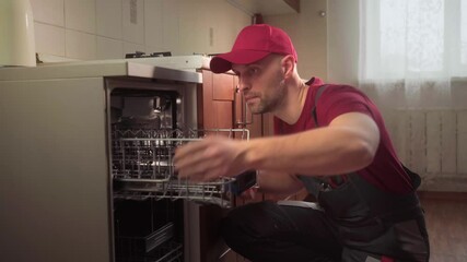 Professional maintenance worker wearing red uniform carefully diagnosing and repairing dishwasher inside residential kitchen, using specialized tools and expertise