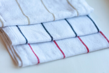 Neatly folded, three white tea towels with thin stripes in neutral tones of gold, dark blue, and red sit on a white surface.