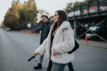 Naklejka premium A fashionable woman wearing a white fur jacket is seen crossing a city road during a slightly overcast day, complemented by urban surroundings and casual attire, embodying style and confidence.