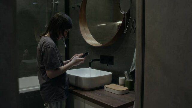 Medium full side shot of young girl standing by sink in bathroom, brushing teeth while scrolling social media, typing messages on smartphone, communicating with online bullies or controlling boyfriend
