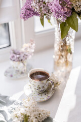 porcelain teacup with freshly brewed coffee, featuring delicate foam and visible steam, placed beside a glass vase with a vibrant lilac bouquet. A cozy and aromatic scene of elegance and nature.