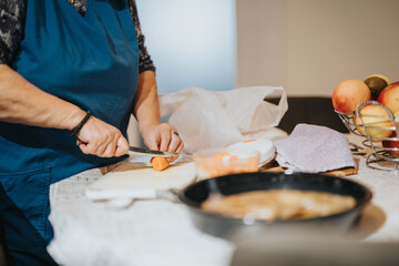 An older woman is seen chopping vegetables on a cutting board, surrounded by apples and cooking essentials on a table.
