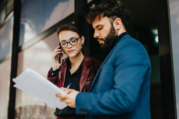 Two business professionals collaborate outdoors, reviewing documents and engaged in a mobile phone...