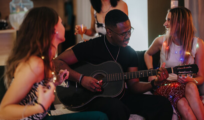 Obraz premium A joyful gathering of friends with a man playing guitar, surrounded by warm lights, sharing positive energy while celebrating an event in a relaxed and festive atmosphere.
