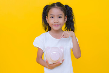 Smiling girl pointing at piggy bank on yellow background