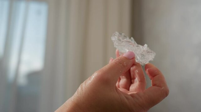 Close up of hand holding clear rubber-like soluble material with water droplets, pink polished nails, natural daylight shining on translucent texture, against soft blurred indoor background