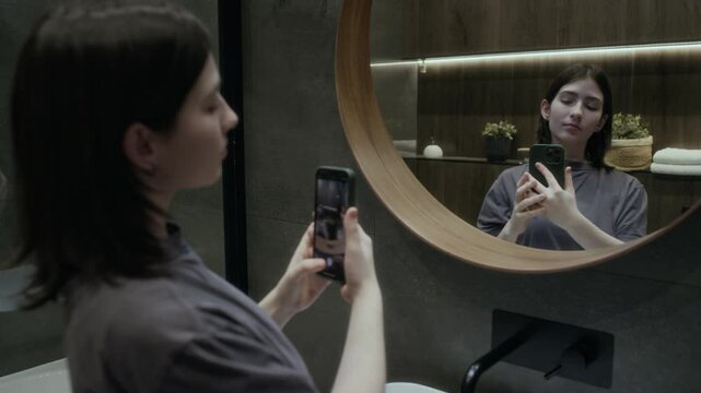 Medium closeup of young Caucasian girl preening in front of mirror, taking selfies with contrived smiles on smartphone for social media, trying various angles and face expressions