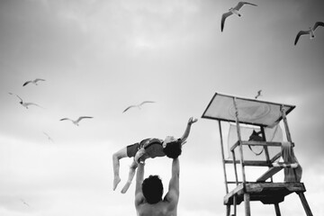 Black and white photo of father lifting child at the beach near lifeguard tower with seagulls...