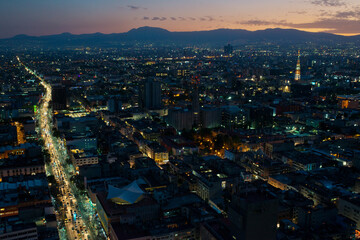Obraz premium Aerial View of Mexico City at Dusk with Illuminated Streets and Distant Mountains.