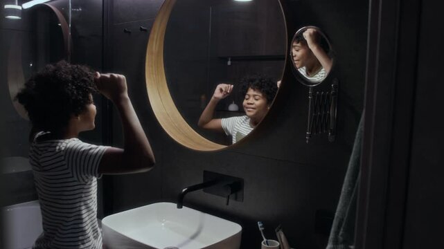 Medium rear shot of young adolescent African American boy with curly hair standing alone in bathroom in front of mirror, flexing biceps, touching strong arm muscles, looking at reflection and smiling