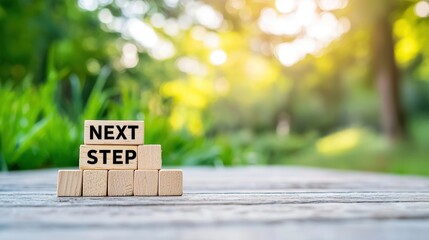 Wooden blocks spelling "NEXT STEP" outdoors on a wooden surface with a bright, green, and sunny background.