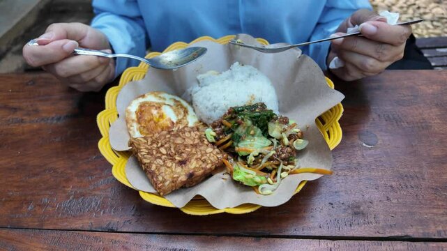 An Indonesian woman eating sego Pecel, the traditional dish from Java, Indonesia, consisting of boiled vegetables, peanut sauce, and side dishes.