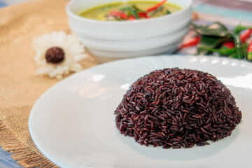 Close up of a mound of cooked black rice on a white plate with Thai green curry in the background, Healthy Asian Food.