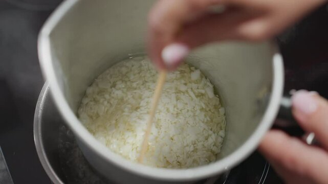 Housewife stirs melting candle wax with stick inside metal bowl placed over steaming pot using double boiling method on a gas cooker, demonstrating homemade candle craft with focus on technique