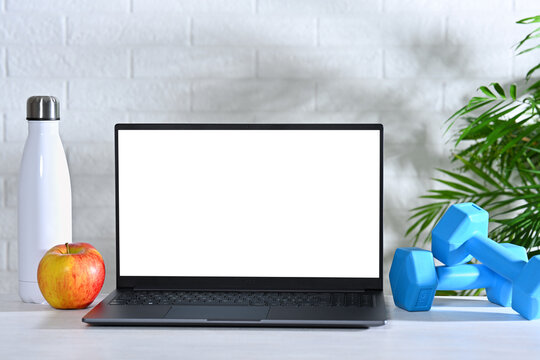 Laptop with white screen on table with dumbbells, bottle and apple