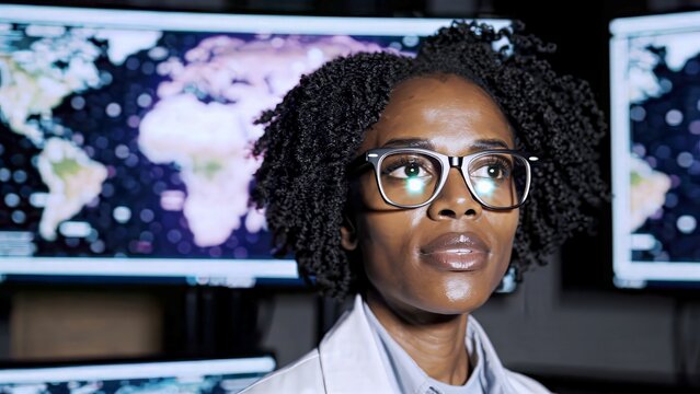 Scientist wearing lab coat and glasses analyzing data reflected in her glasses on multiple computer screens showing world map in a dark room