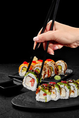 Woman using chopsticks picking sushi rolls from plate on black background