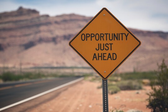 Orange diamond-shaped road sign against blurred desert landscape, displaying "Opportunity Just Ahead," conveying optimism and anticipation for future success