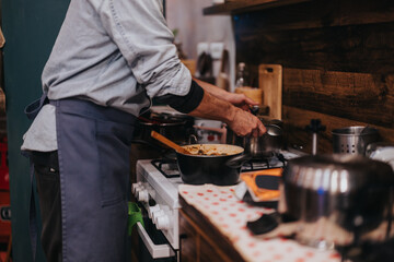 A man wearing a blue apron cooks in a warm, rustic kitchen setting, using pots on a stove. The countertop features various utensils and tools, adding to the domestic atmosphere.
