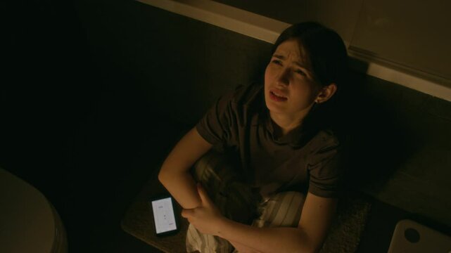 Top view of young Caucasian girl with mental breakdown due to cyberbullying sitting huddled up on floor in dimly lit bathroom and sobbing bitterly. Smartphone with abusive messages lying beside