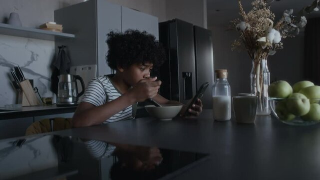 Medium full shot of teenage African American boy sitting at table, eating cornflakes with milk for breakfast, opening abusive message in group chat on smartphone, dropping spoon, staring in dismay