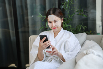 Asian woman in white bathrobe relaxes on sofa while using her smartphone, with facial sheet masks...