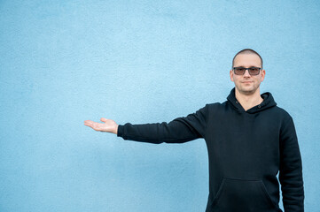 Stylish guy in glasses and black sweatshirt isolated on plain bright blue background, pointing with...