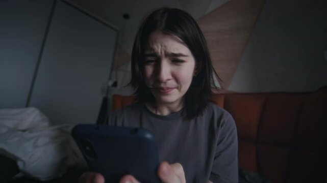 Dutch angle medium close-up of dismayed teenage girl sitting in messy dark bedroom alone, reading vicious messages from online bullies, sobbing, shouting and screaming in anguish and fury