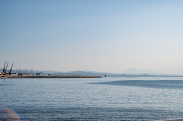 Calm sea with gentle ripples and a stone breakwater extending into the water. Port cranes are visible on the horizon under a clear sky.