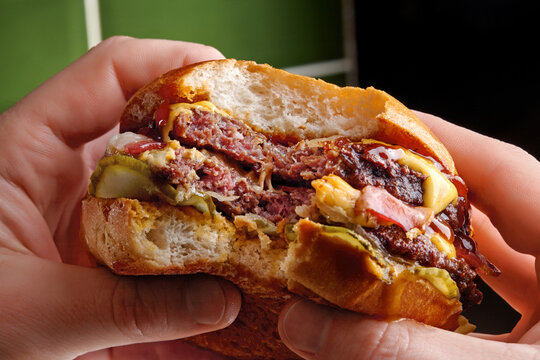 Hands holding a double burger with bacon, pickles, and mustard