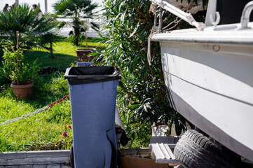 A plastic trash bin stands next to a docked boat near a green lawn and potted plants, close to the beach area.