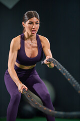 fit woman in purple activewear uses battle ropes enhance strength and endurance in gym setting. intense workout showcases her dedication and effort in dynamic environment.