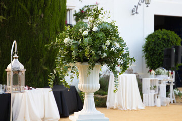 Flower pot with eucalyptus branches and white flowers in the decoration for an outdoor wedding. There are catering tables and lanterns in the decoration of the event.