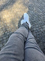 casual snapshot of crossed legs in grey jeans and white sneakers, resting on a sunlit cobblestone...