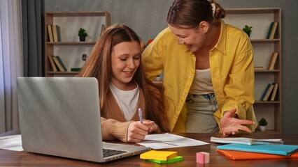 Young teenage girl studying at home, girl sits at desk with laptop and books, doing homework writing test, mother explaining and helping with task, positive expression.