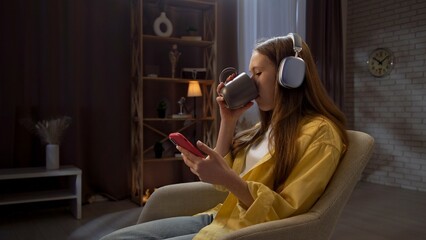 Young teenager girl spending evening time at home, girl sits in chair in the living room drinking...