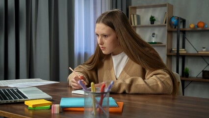 Young teenage girl studying at home, girl sits at desk with laptop and books, doing homework drawing in notebook with liner and pencil, looking at screen.