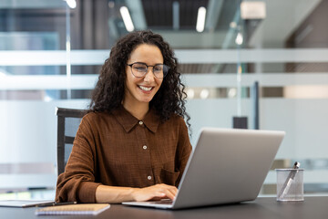 A cheerful woman with glasses works at a desk in a modern office, focused on her laptop with a big...