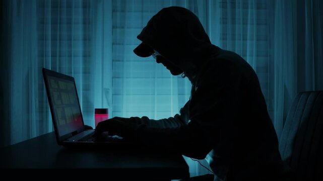 Silhouette of a hacker in hoodie typing on a laptop with code on screen in a dark room. Cybercrime and data breach concept.