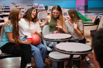 Four female friends are sitting, talking, and laughing together at a bowling alley, enjoying their time