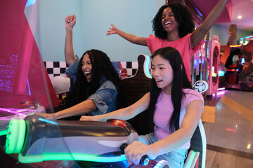 Group of diverse young women having fun while playing arcade racing game in entertainment center