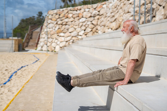 Active senior man performing seated leg lift exercise on concrete steps outdoors, demonstrating strength, balance, and a healthy lifestyle. 